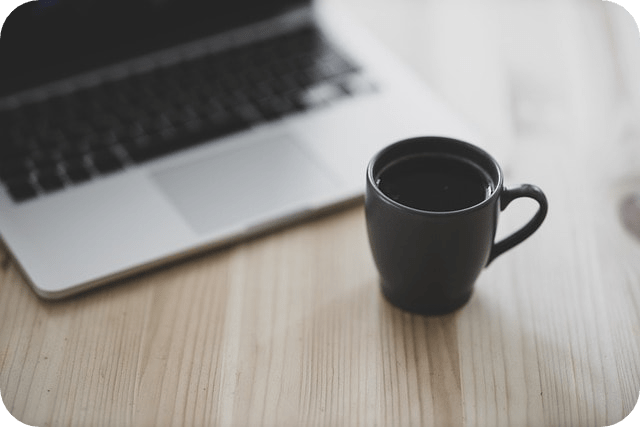 A black coffee mug placed on a wooden table next to an open laptop, suggesting a work or study setup with a casual vibe.
