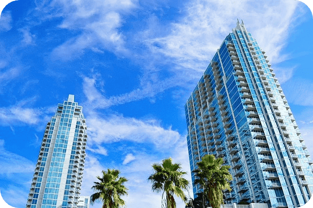 Two high-rise buildings with glass facades stand tall against a bright, blue sky dotted with clouds.